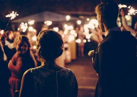 A couple stands facing a crowd, with sparklers lighting up the night, suggesting a celebratory or wedding event.